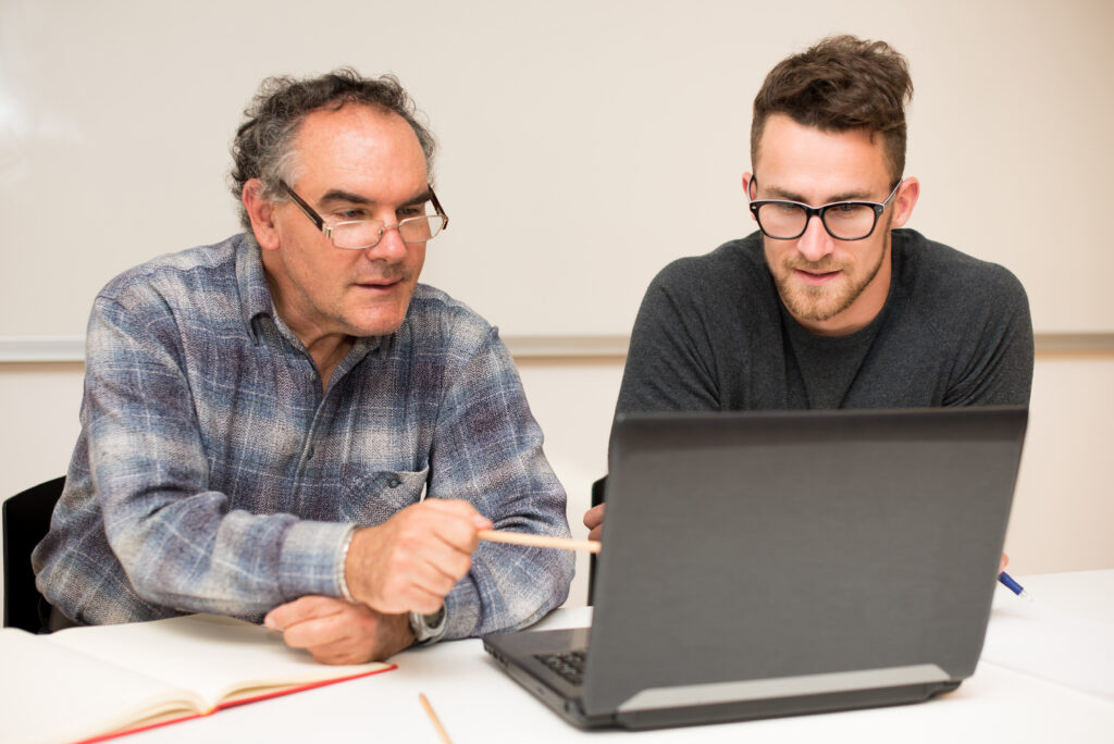 SBA-SCORE-Program-Business-Mentor An older man teaching a younger man by pointing to something on a computer.