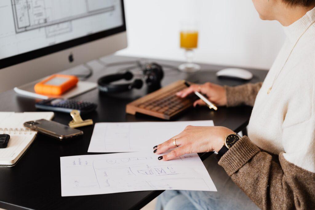 An employee is hard at work, looking at a computer screen with a hand on the papers on the desk in front of them.