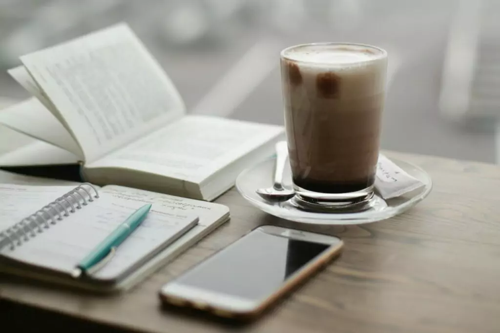 A glass of coffee, a book, a notebook with a pen, and a cell phone sit on the table at a coffee shop.