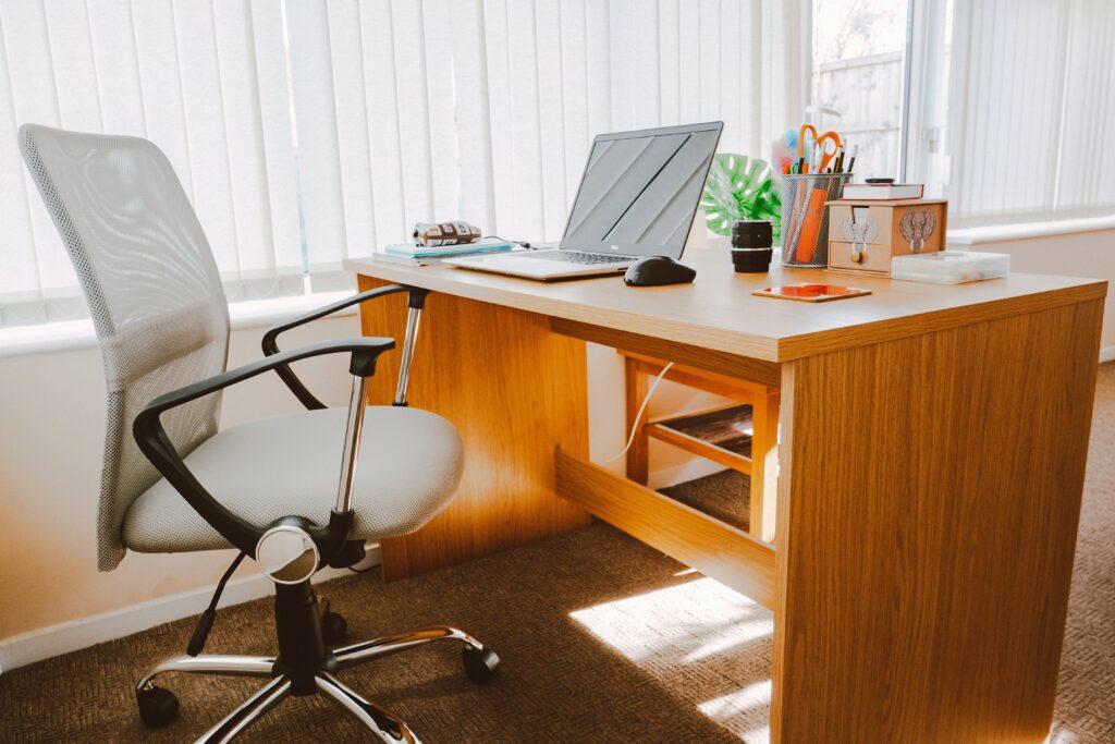 An employee's desk next to a window, with a laptop, computer mouse, and other things on it, along with an empty desk chair.
