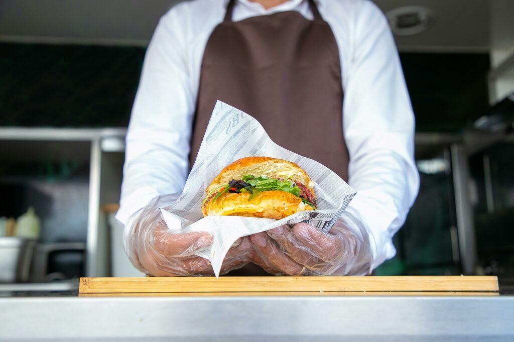 The owner of a restaurant, wearing gloves and an apron, holds a chicken sandwich in front of them to display it.