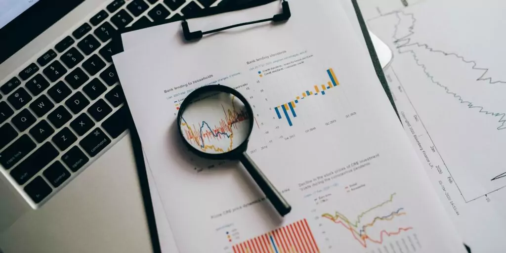A laptop and papers showing business finances, along with a magnifying glass. This signifies close analysis of business finance data.